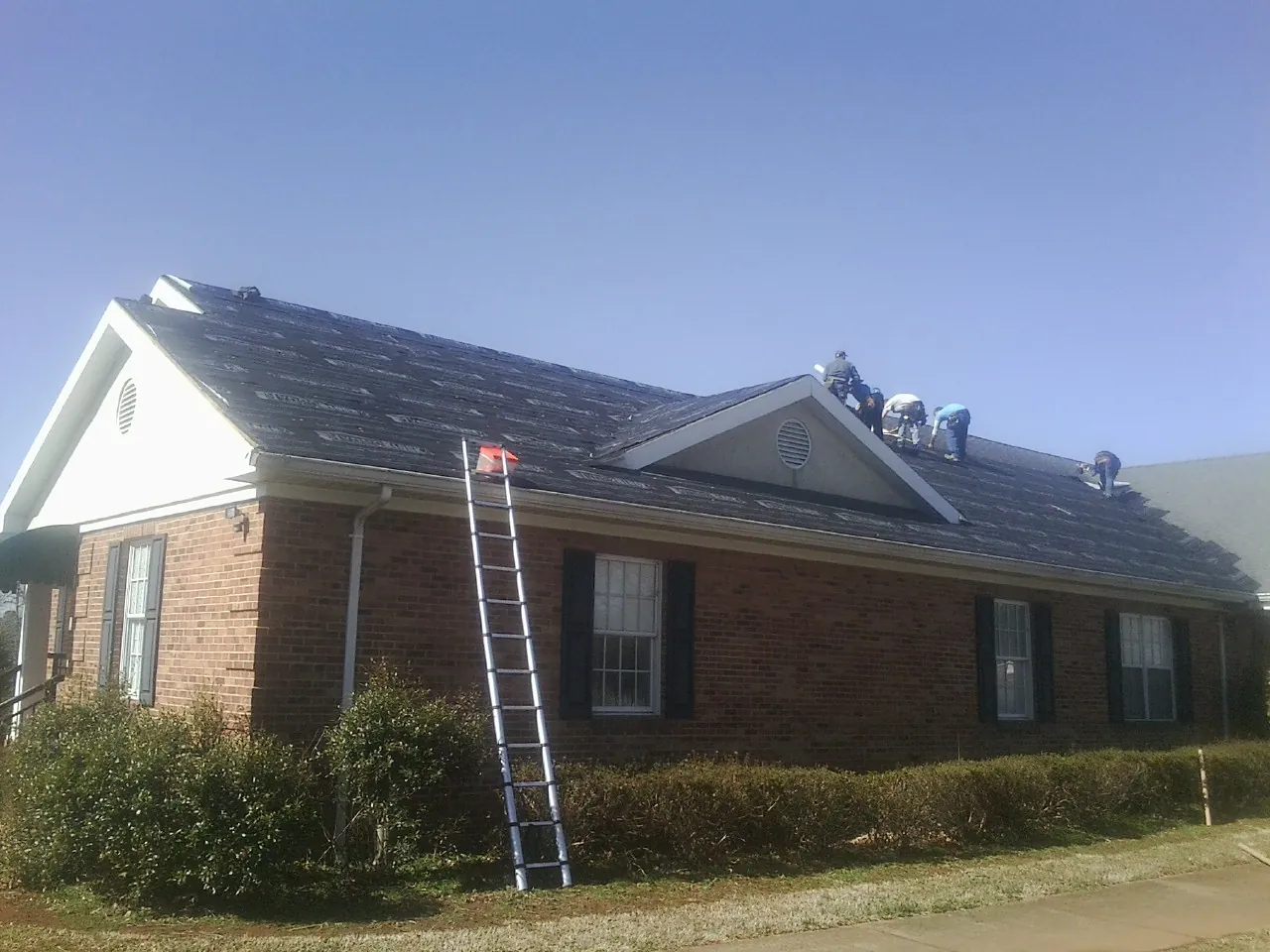 Crew tearing shingles off a brick ranch roof with a ladder set — Emergency Roof Repair work by our crew in Lostant