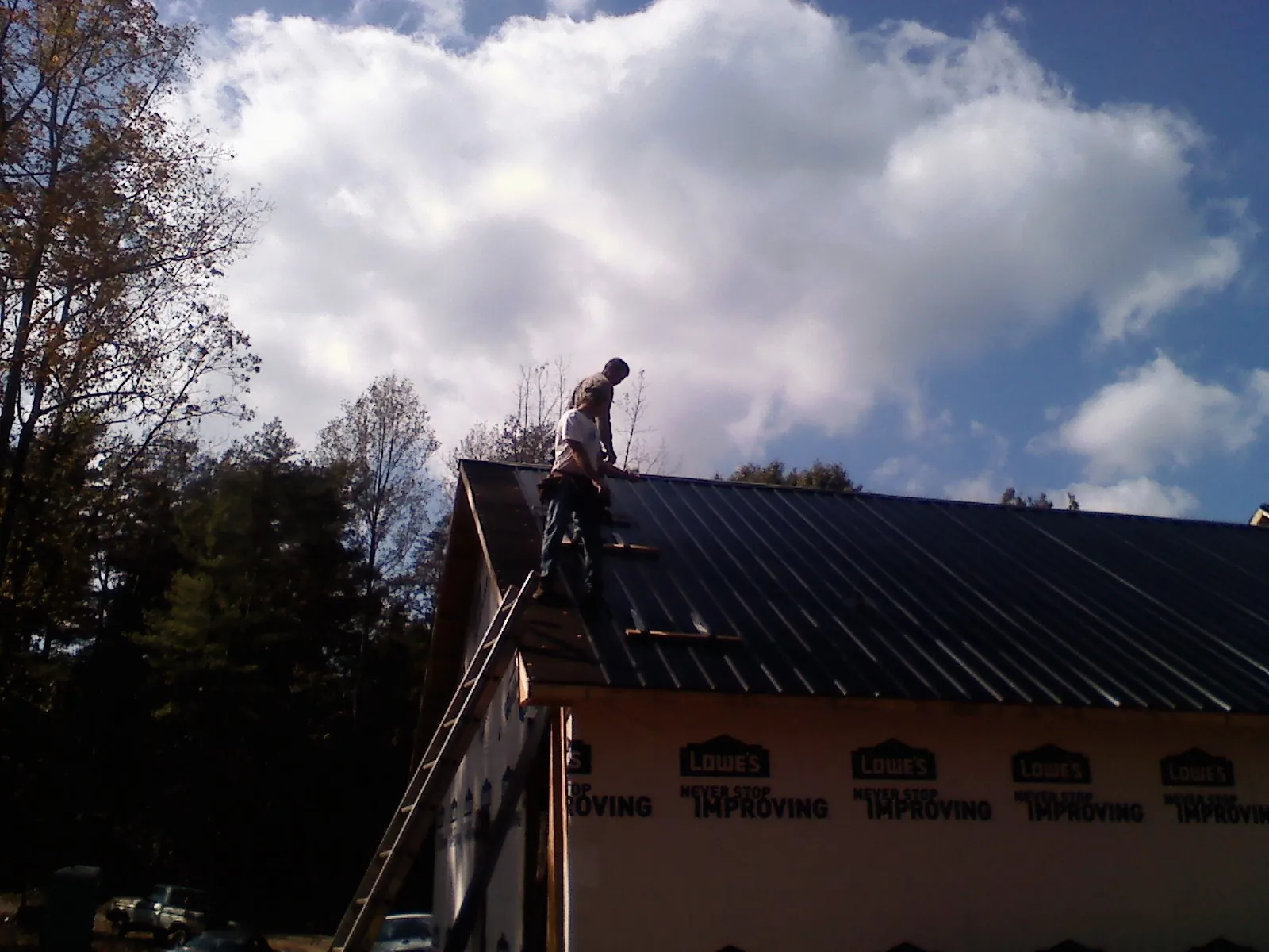 Roofer fastening dark metal panels high against a cloudy sky, from a Roof Inspection job in Lostant